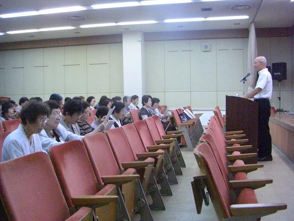 赤い椅子に20名ほどの女性たちが座っていて、その前の教壇に市長が立って話をしている写真